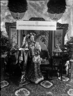 Putting a flower in her Manchu-style coiffure.  Cixi took great care of her appearance.  She designed her clothes and jewellery and supervised the making of cosmetics such as rouge, perfume and soap.  In the background, apples from her orchard were on display for their subtle fragrance.